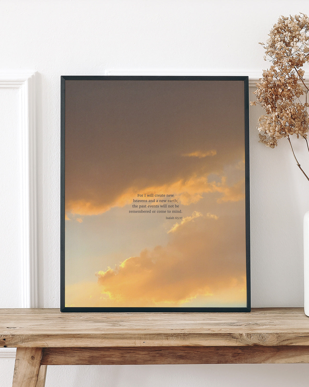 A wooden bench against a white wall with decorative molding, holding a framed picture displaying scripture art from nature with a vibrant sunset sky and a quote from Isaiah 65:17 by Heaven & Nature Co. Next to it, a white vase contains dried beige hydrangeas.