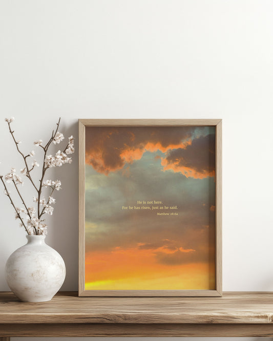 Framed Scripture art print with Matthew 28:6a on an Easter-colored sky. The print is on a wooden surface with a vase and branches.