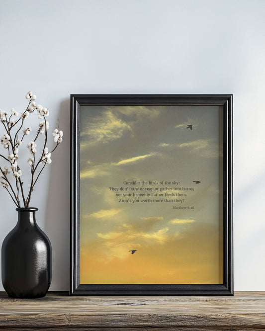 Framed Scripture art print with Matthew 6:26 on a sunset summer sky with three black birds flying. The print is leaning on a wall next to a vase with branches.