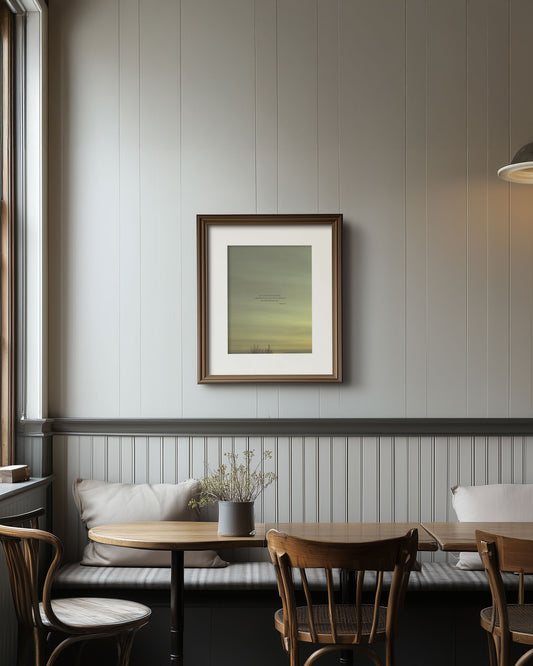 Framed Scripture art print with Micah 7:7 on a greenish yellow sky with the tops of bare trees at on the bottom. The print is on a wall in a room with wooden tables and chairs.