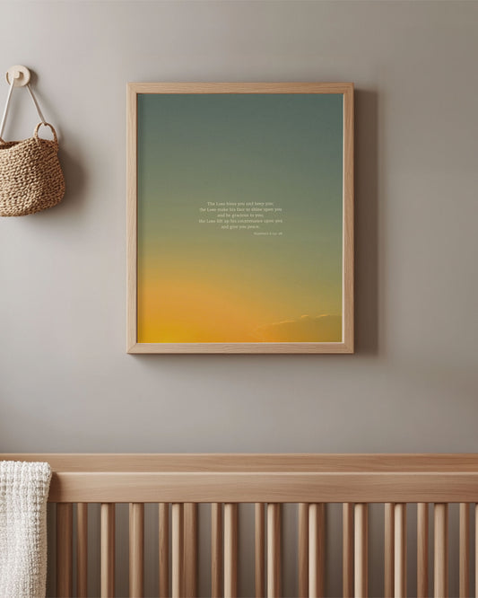 Framed Scripture art print with Numbers 6:24-26 (ESV) on top of a gradient orange and blue sky hanging above a crib in a room.