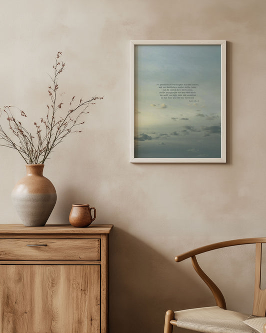 Framed Scripture art print with Psalm 108:4-6 on a sky with a gray-blue gradient and soft sunlight illuminating small clouds. The print is on a wall above a wooden cabinet with decorative items.