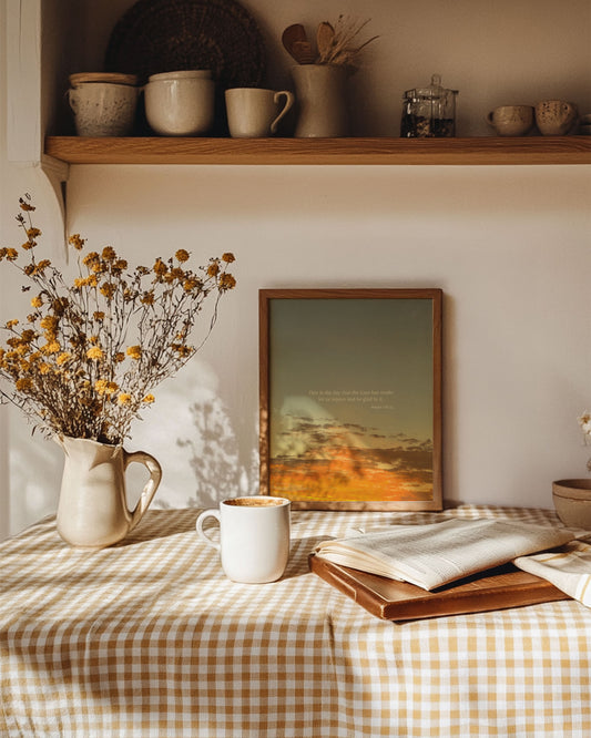 Cozy home setting with a table, mug, book, and framed Scripture art print of Psalm 118:24 with the sun overcoming dark clouds. The print is sitting on a checkered tablecloth.