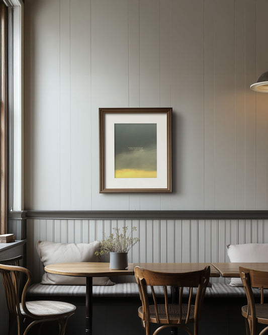 Framed Scripture art print with Psalm 124:8 on a glowing yellow to dark-grey gradient cloudy sky. The print is on a wall in a room with wooden tables and chairs.