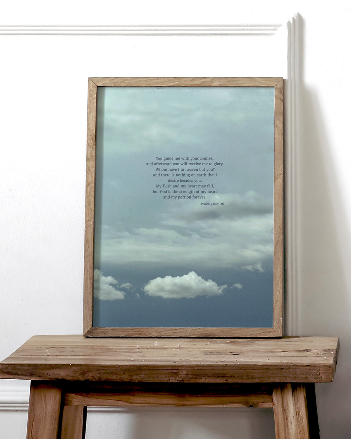 A wooden side table holding a framed picture displaying scripture art from nature with a cloudy sky and a quote from Psalm 73:24-26 by Heaven & Nature Co.