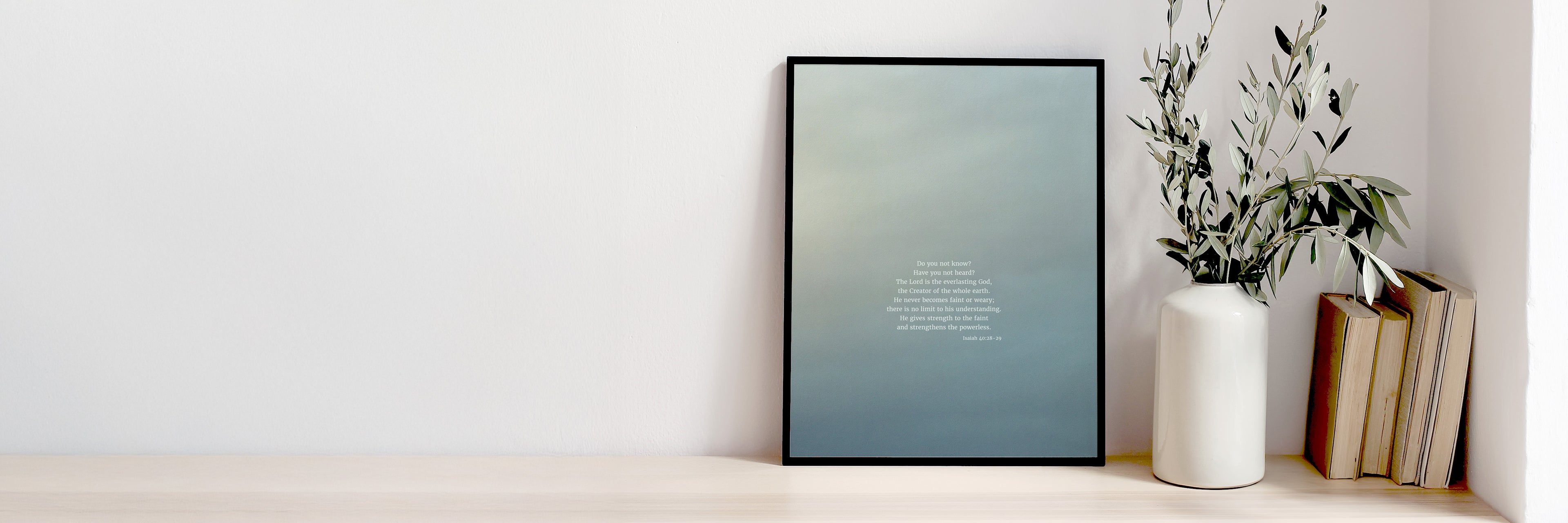 A minimalist shelf with a framed picture displaying scripture art from nature of Isaiah 40:28-29 by Heaven & Nature Co., a white vase with olive branches, and a stack of old books on the right side, set against a plain wall.