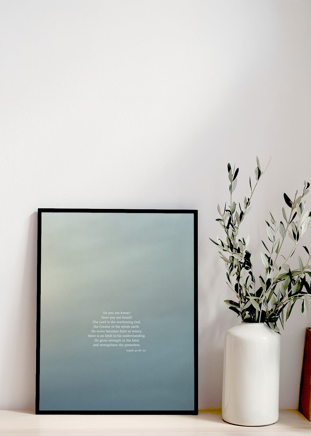 A minimalist shelf with a framed picture displaying scripture art from nature of Isaiah 40:28-29 by Heaven & Nature Co., a white vase with olive branches, and a stack of old books on the right side, set against a plain wall.