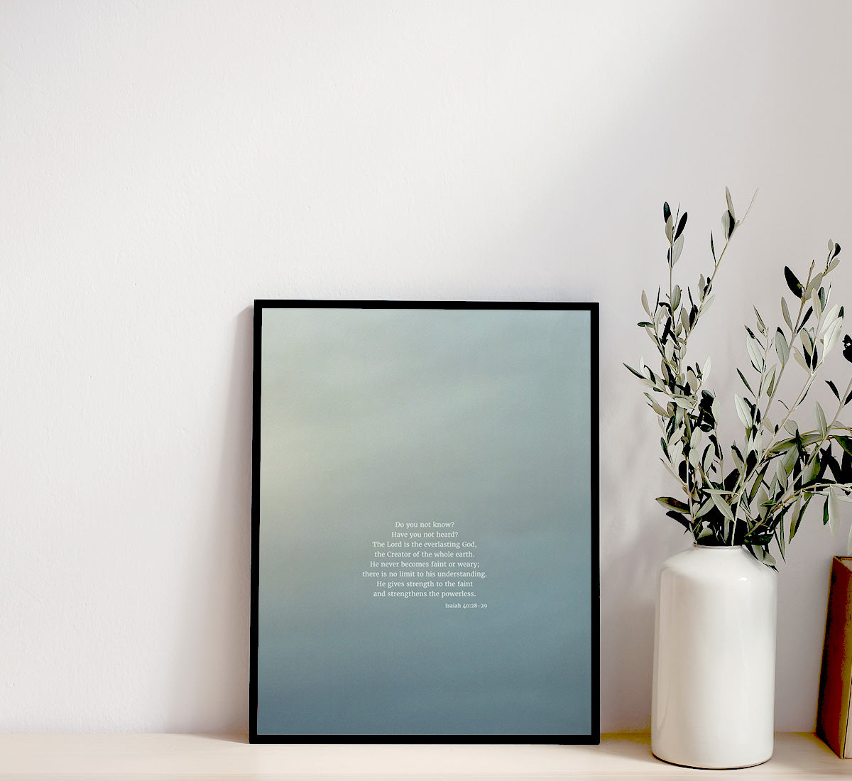 A minimalist shelf with a framed picture displaying scripture art from nature of Isaiah 40:28-29 by Heaven & Nature Co., a white vase with olive branches, and a stack of old books on the right side, set against a plain wall.
