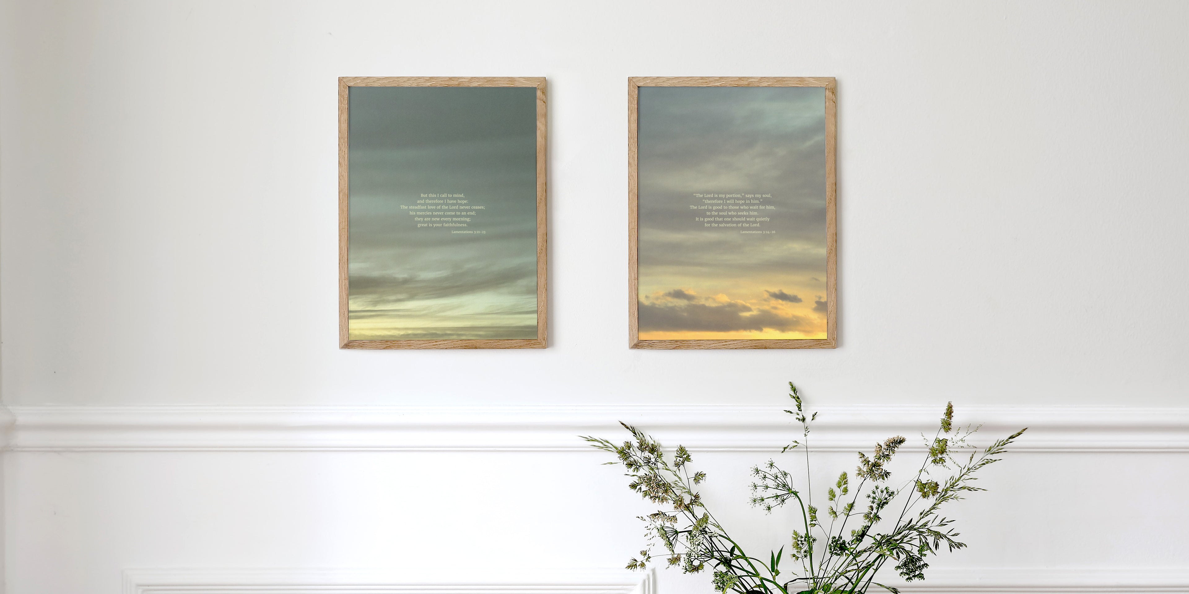 Two framed pictures displaying scripture art from nature of sky backgrounds and quotes from Lamentations 3:21-23 and 3:24-26 by Heaven & Nature Co., hung on a white wall with a decorative molding. Below, a vase with green and white grasses adds a natural touch.