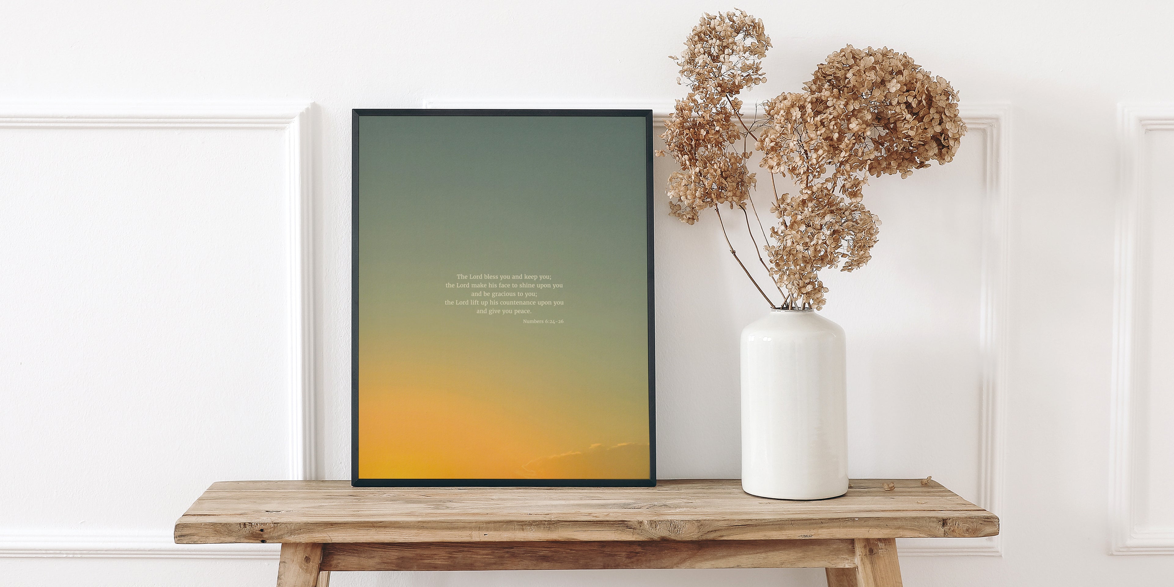 A wooden bench against a white wall with decorative molding, holding a framed picture displaying scripture art from nature with a gradient sky background and a quote from Numbers 6:24-26 by Heaven & Nature Co. Next to it, a white vase contains dried beige hydrangeas.